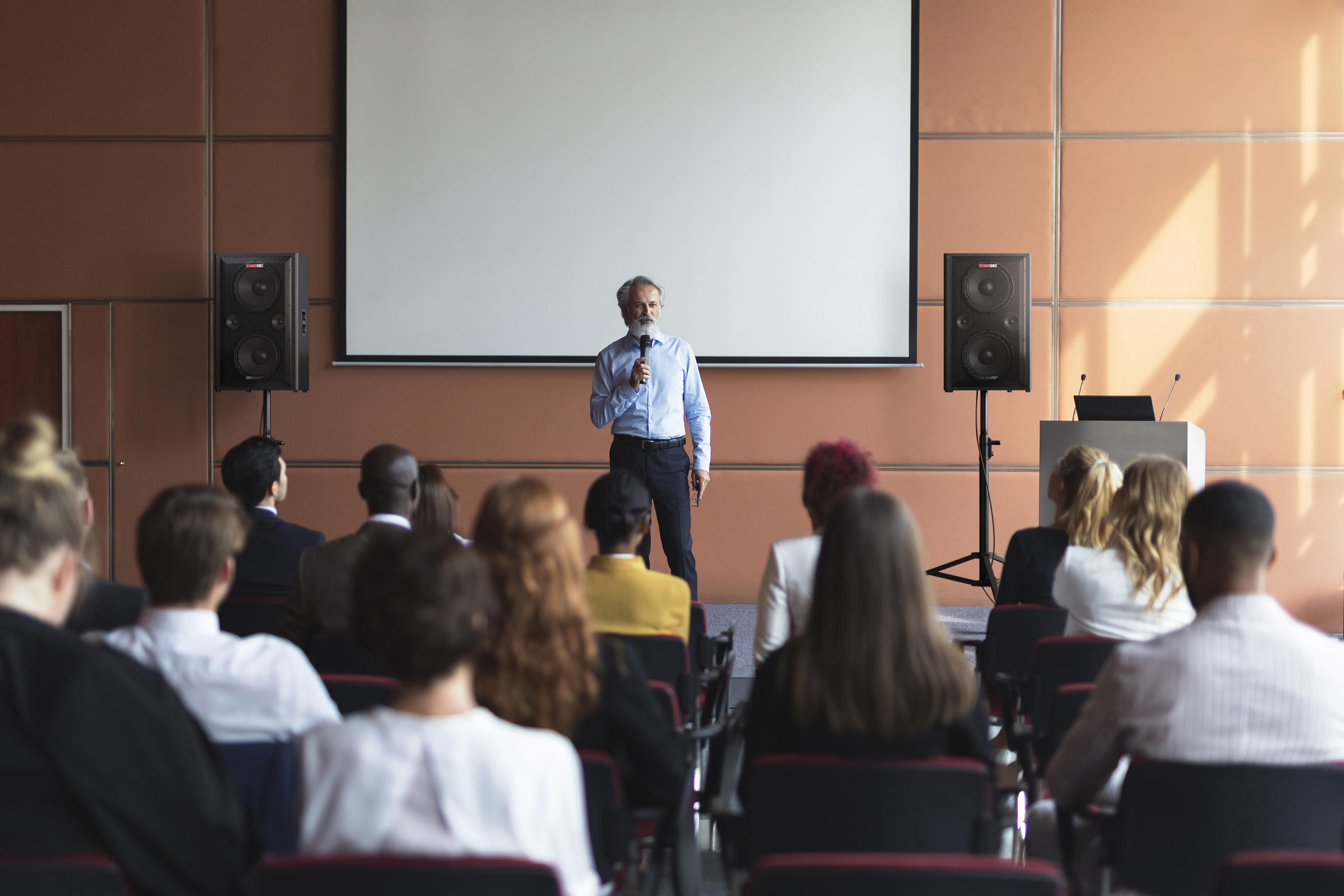 Un uomo parla a una conferenza. Pubblico seduto di fronte a lui. Altoparlanti su entrambi i lati.