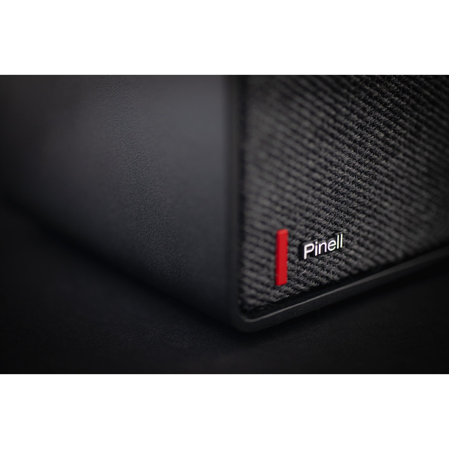 Close-up of black speaker, with a textured fabric covering. Red Pinell logo is visible.