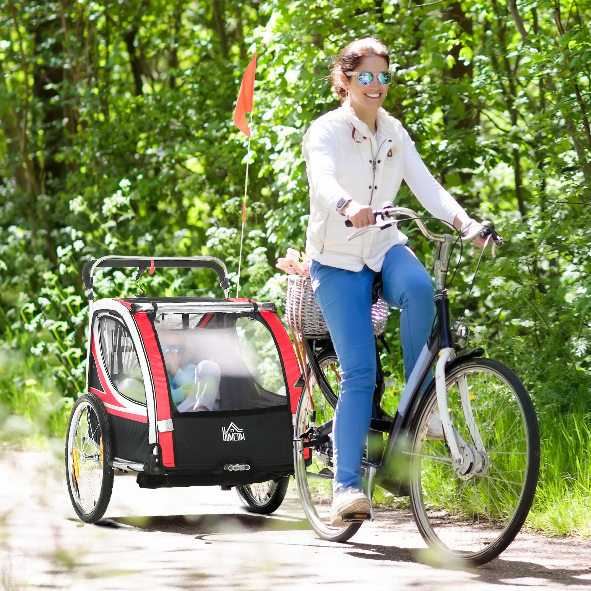 Donna in bicicletta, che tira un rimorchio per bambini. Il rimorchio ha parti rosse, nere e bianche. Sfondo di alberi verdi e sentiero.
