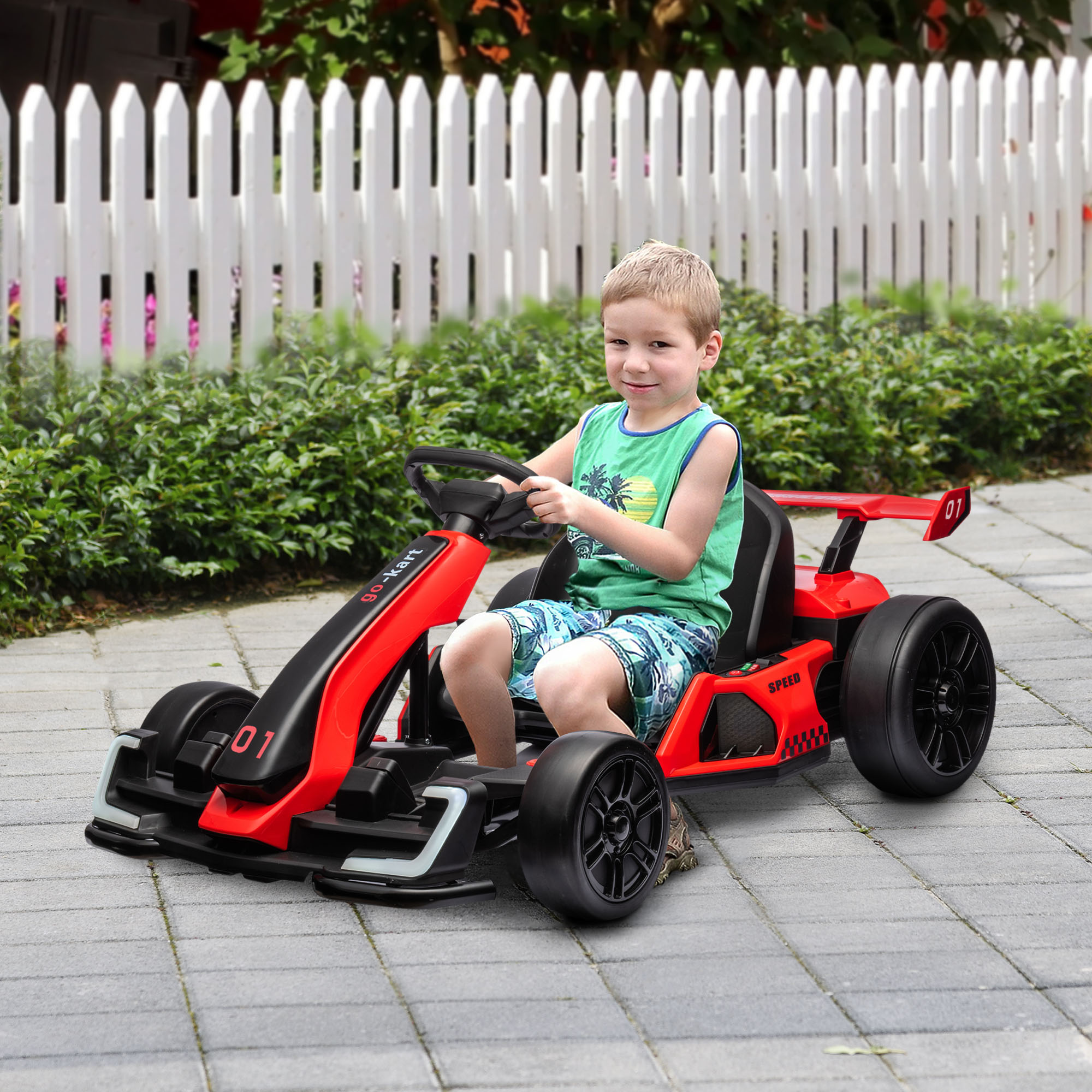 Un bambino in camicia verde e pantaloncini blu è seduto su un go-kart rosso, sorridendo. Recinzione bianca sullo sfondo.