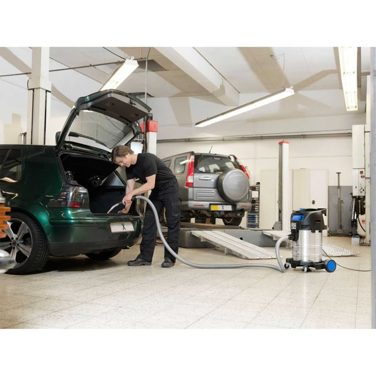 Ein Mann saugt einen Kofferraum in einer Garage, ein Staubsauger steht daneben. Ein anderes Auto ist im Hintergrund.