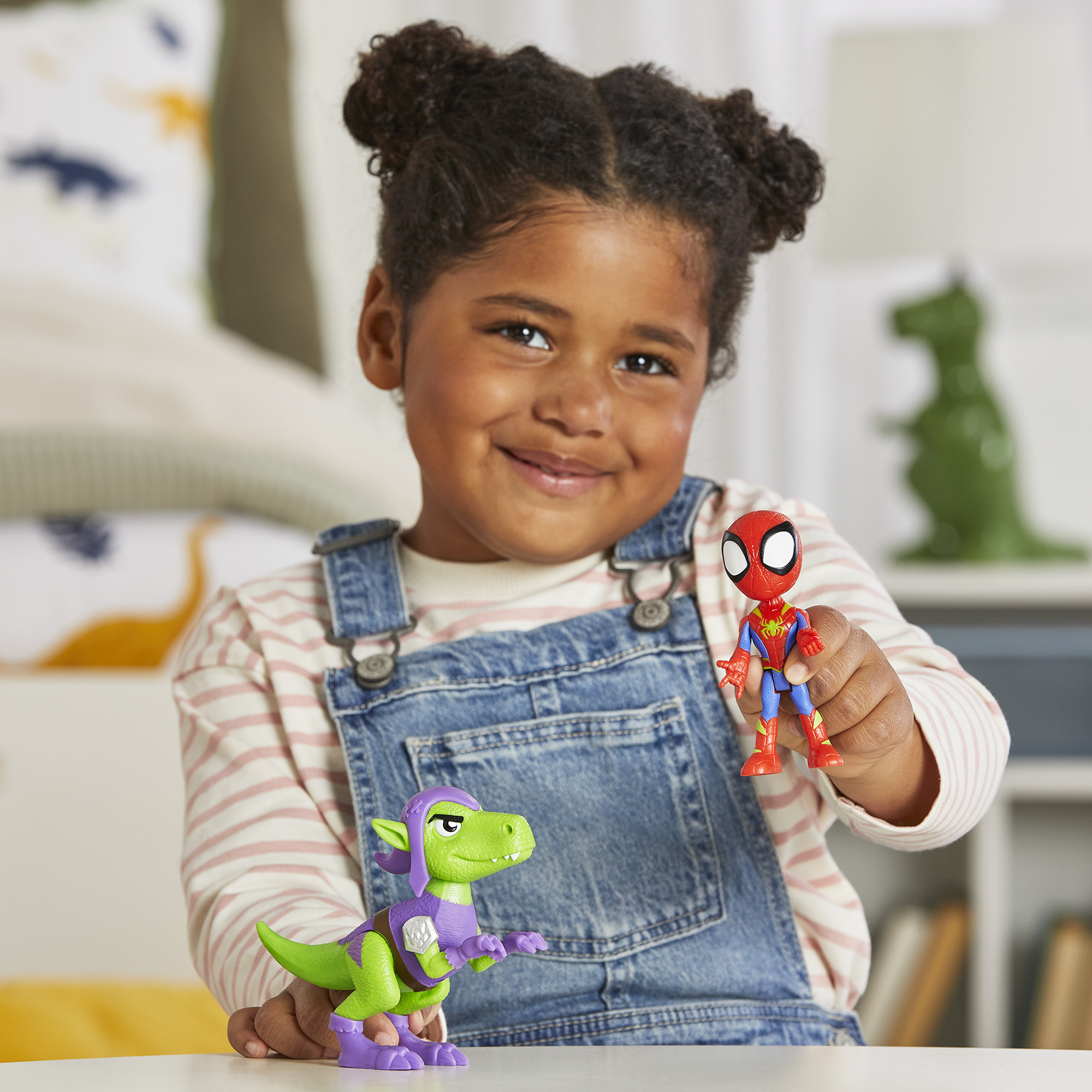 Una niña sonriendo y sosteniendo un Spiderman y un juguete de dinosaurio verde. El fondo es un dormitorio.