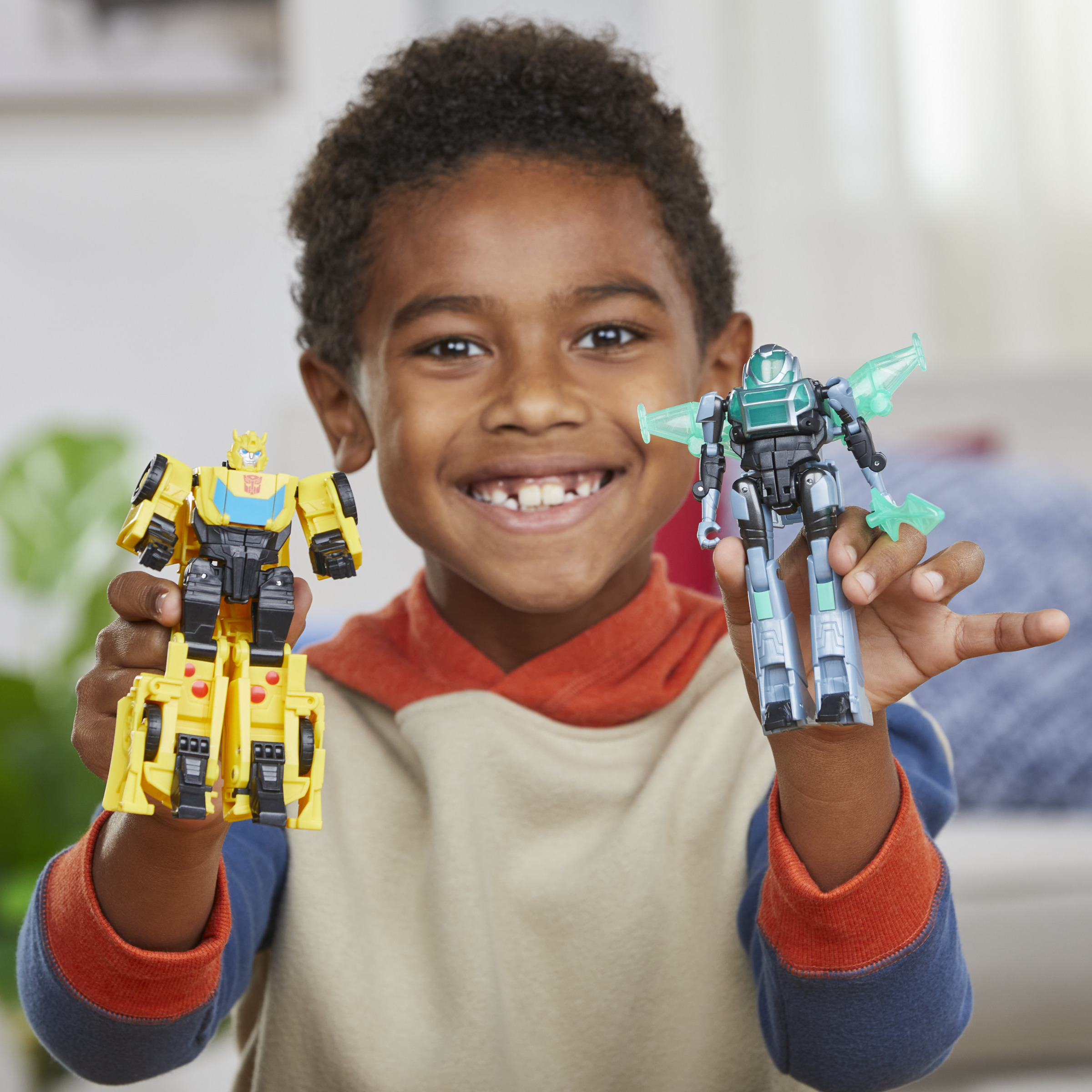 Un niño sonriente sosteniendo dos juguetes Transformers, un coche-robot amarillo y un jet-robot verde.