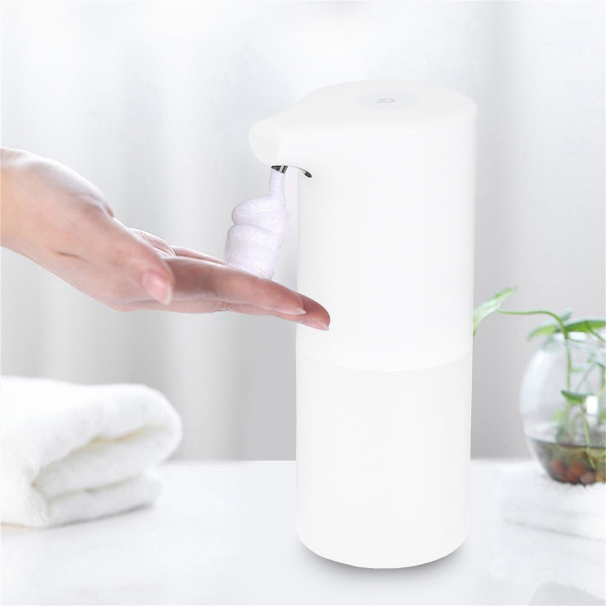 Eine Hand streckt sich aus, um Schaum aus einem weißen automatischen Seifenspender zu empfangen. Handtuch und Pflanze sind in der Nähe.