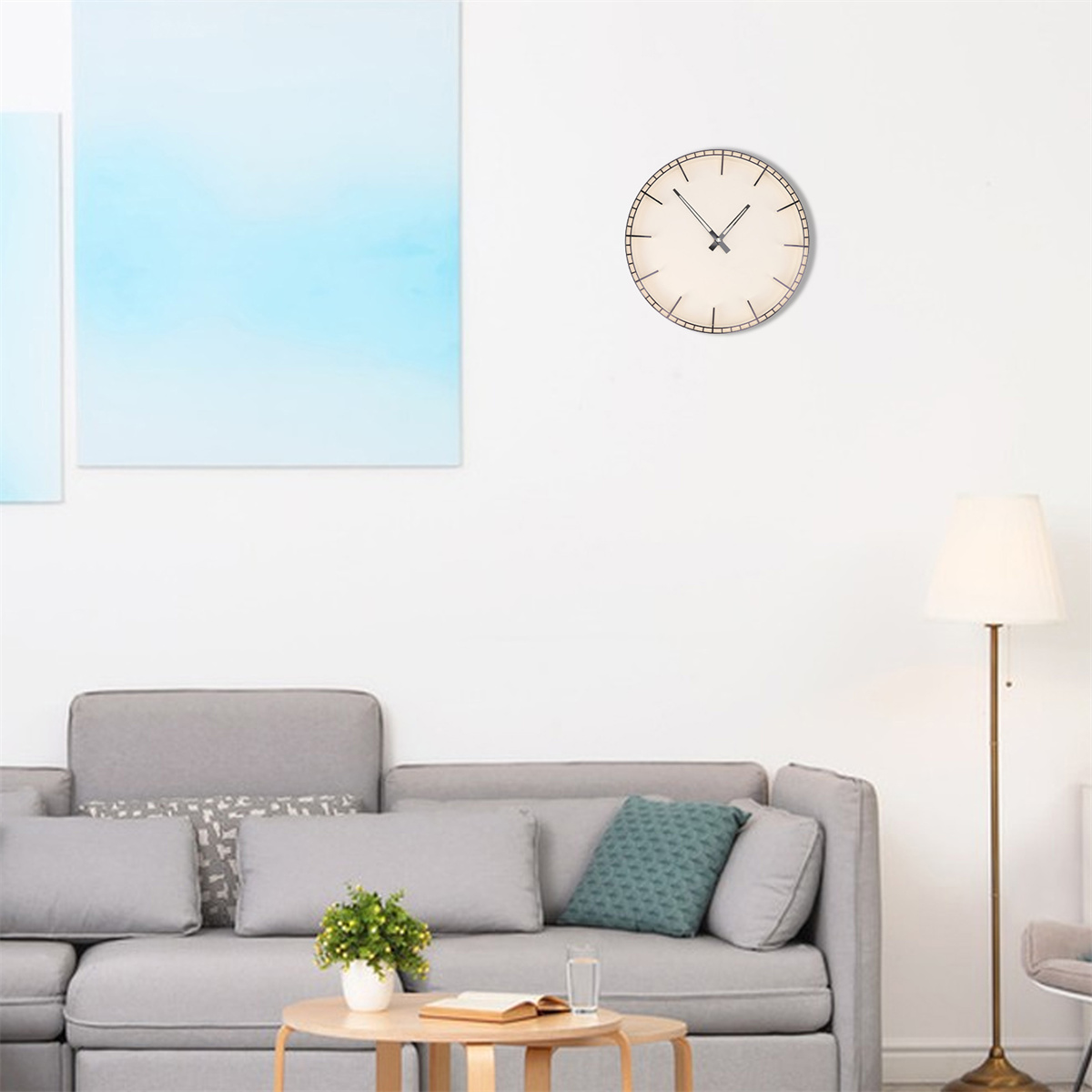 Eine Uhr an einer weißen Wand über einem grauen Sofa mit Kissen. Eine Lampe und Gemälde sind ebenfalls im Raum.