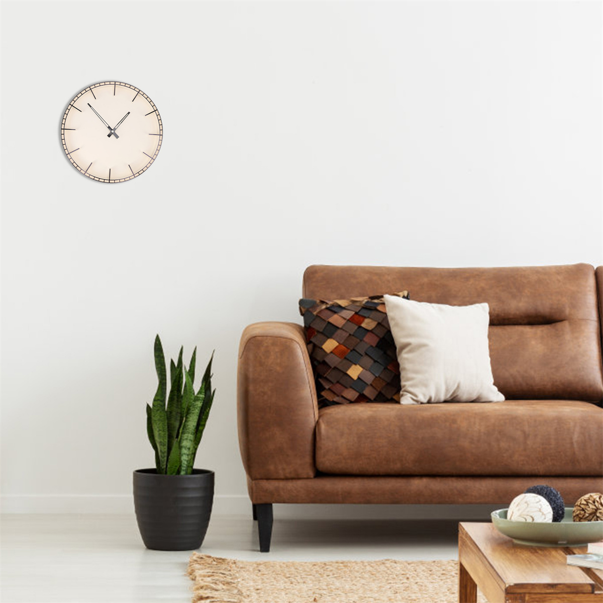 Ein braunes Ledersofa, Uhr an der Wand, eine Pflanze und Kissen auf dem Sofa und ein kleiner Tisch.