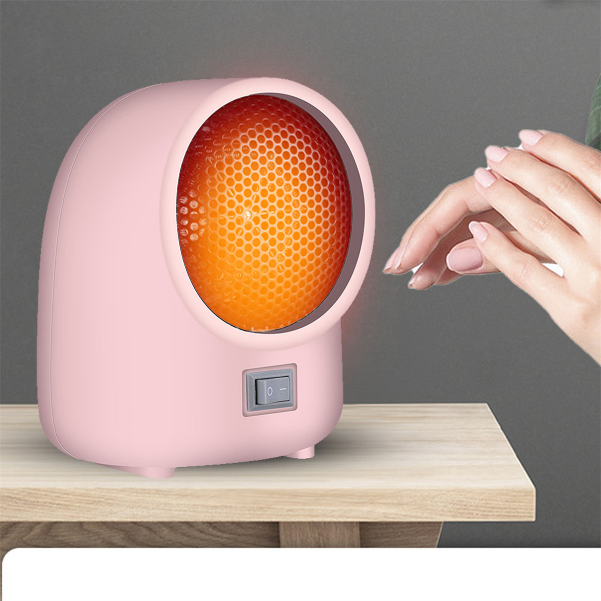 Ein rosa, abgerundeter Heizlüfter mit orangefarbenem, glühendem Heizelement. Eine Hand greift auf einer Holzoberfläche danach.