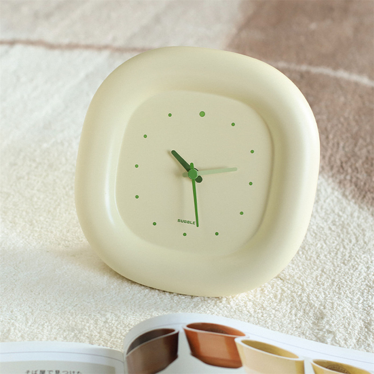 Cremefarbene, quadratische Uhr mit grünen Punkten. Grüne Zeiger und ein verschwommener Hintergrund.