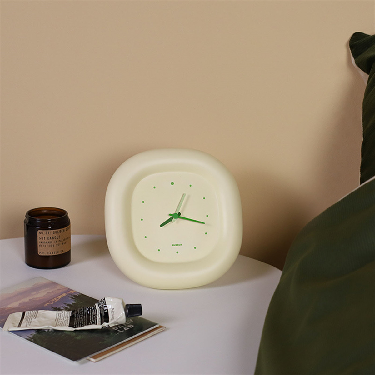 Eine moderne Uhr auf einem weißen Tisch, mit einem Kerzenglas und einer Cremetube. Im Hintergrund ein grünes Kissen.