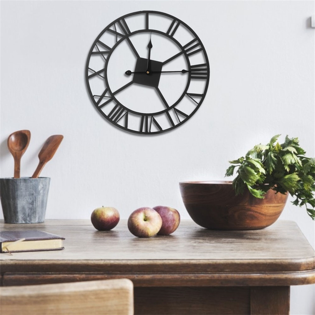 Eine schwarze Wanduhr mit römischen Ziffern an einer weißen Wand. Äpfel und eine Holzschale auf einem Tisch darunter.
