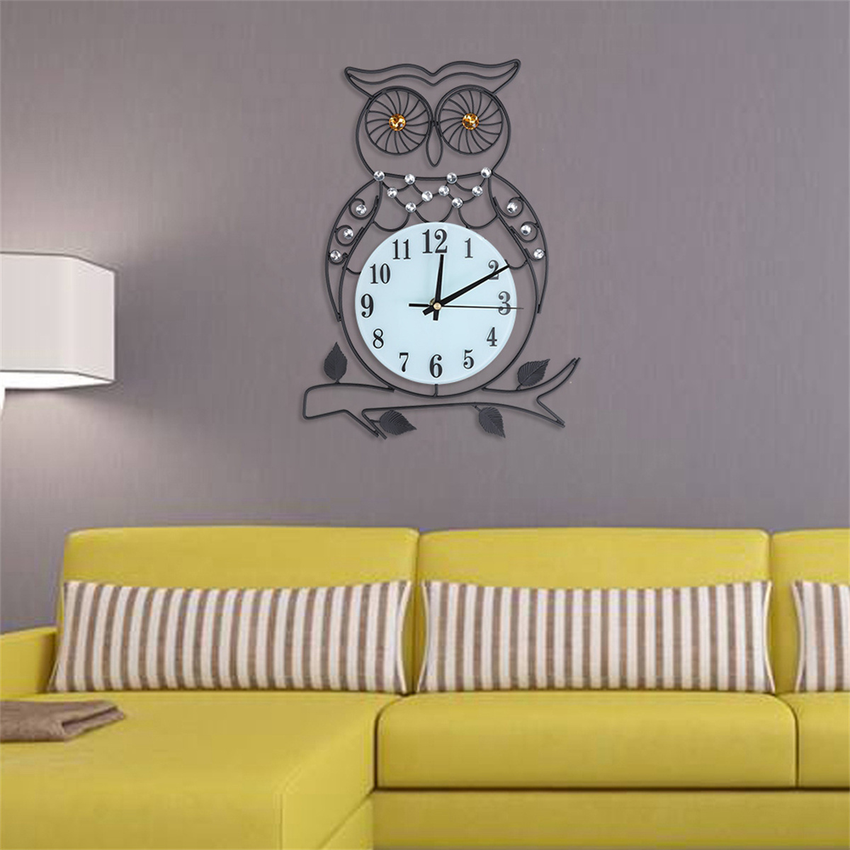 Eulen-Uhr an einer grauen Wand über einem gelben Sofa. Die Uhr hat ein weißes Zifferblatt.