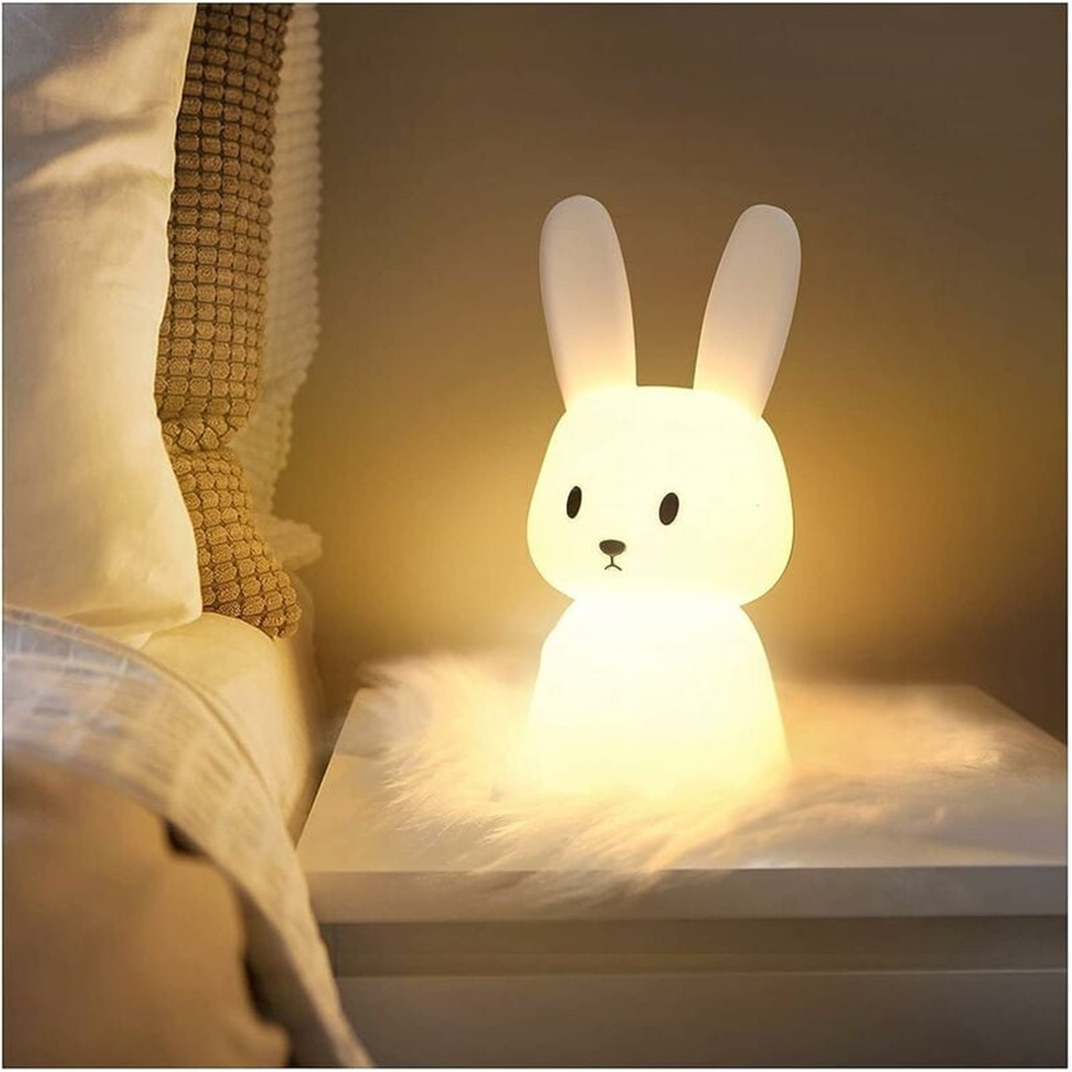 Eine leuchtende weiße Hasenlampe auf einem Nachttisch. Es ist in der Nähe eines Bettes mit weißer Tagesdecke.