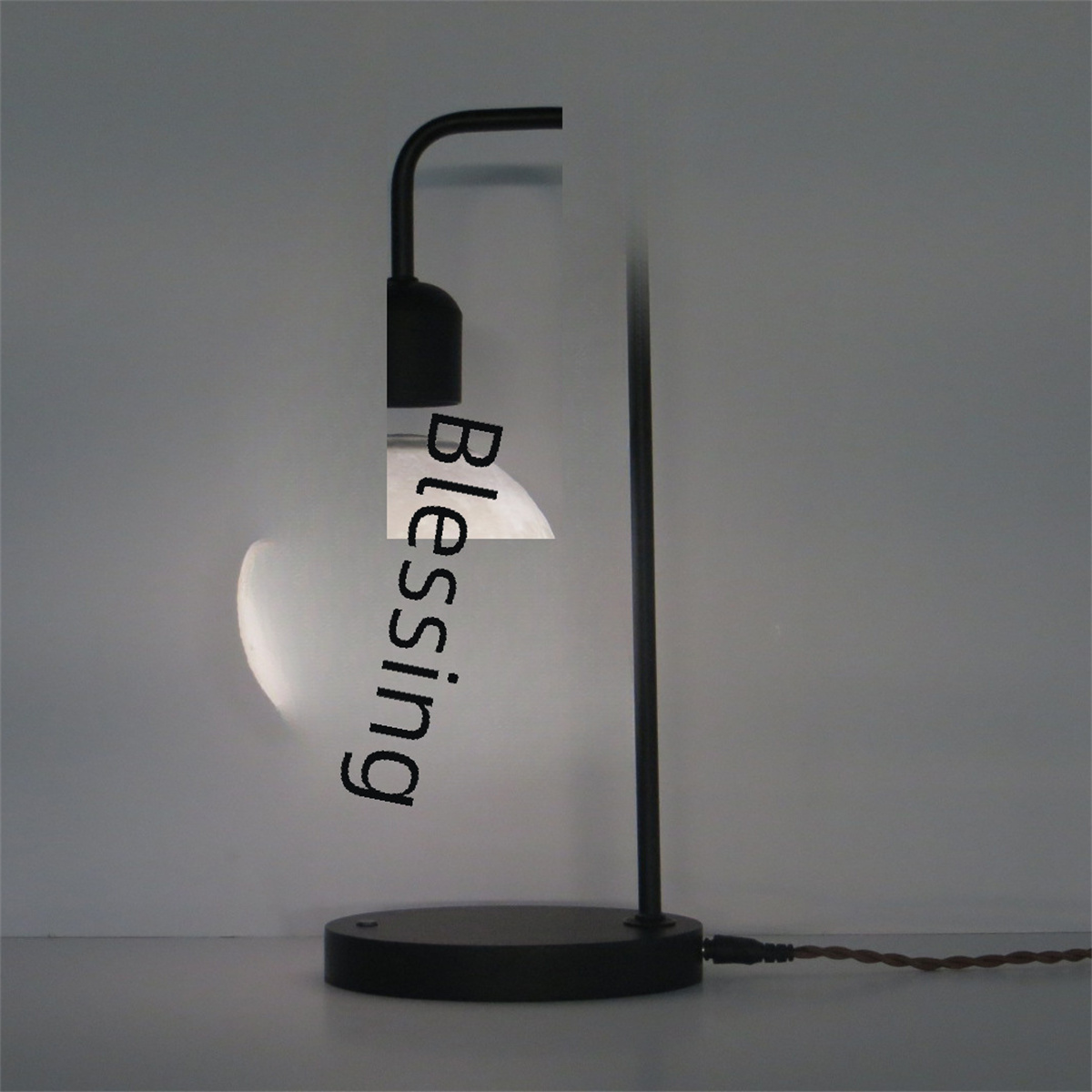 Schreibtischlampe mit runder Glühbirne. Schwarzer gebogener Stiel und Sockel. Auf der Birne steht 'Blessing'.