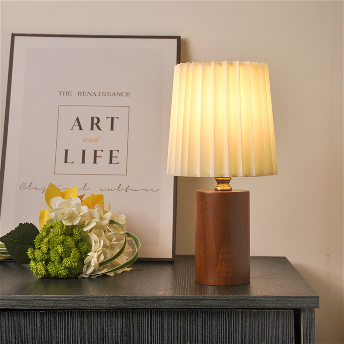 Tischlampe mit plissiertem Schirm. Holzsockel. Blumen und ein gerahmter Kunstdruck links.