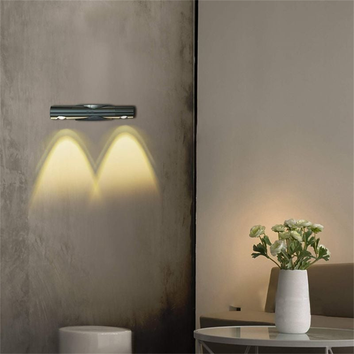 Wandlampe mit zwei Lichtstrahlen, eine weiße Vase mit Blumen auf einem Tisch. Die Wand ist grau.