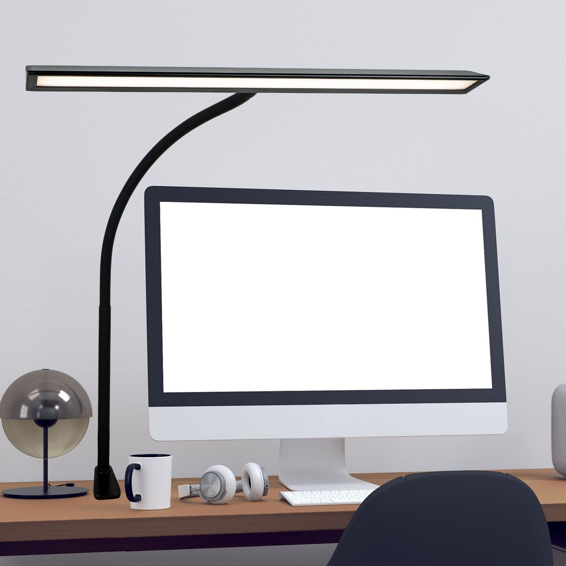 Een moderne bureau-opstelling met een monitor, bureaulamp, mok, koptelefoon en een decoratieve bol.