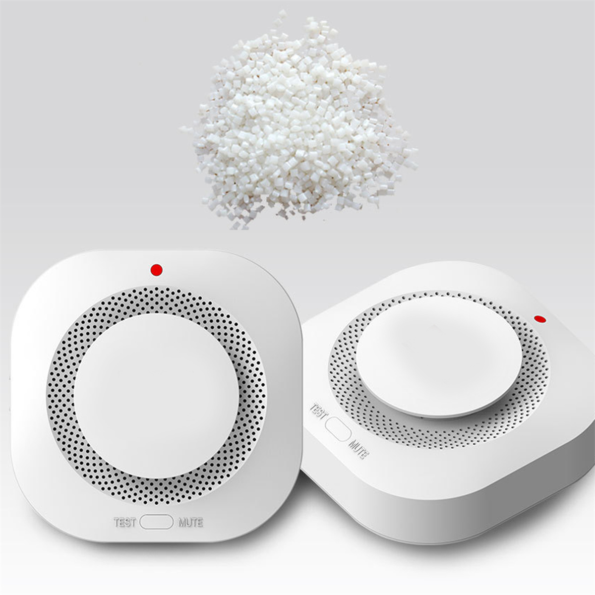 Zwei weiße Rauchmelder mit einem Haufen Plastikpellets im Hintergrund, auf hellgrauem Untergrund.