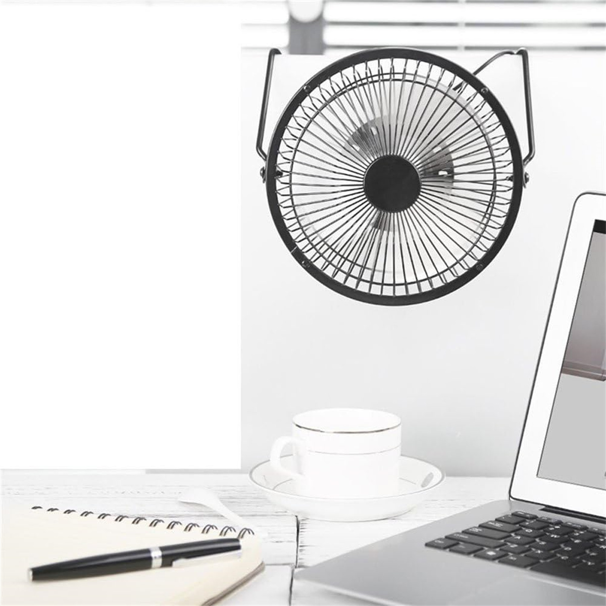 Schwarzer Ventilator, weiße Tasse mit Untertasse, offenes Notizbuch, Stift und Laptop auf weißem Tisch.