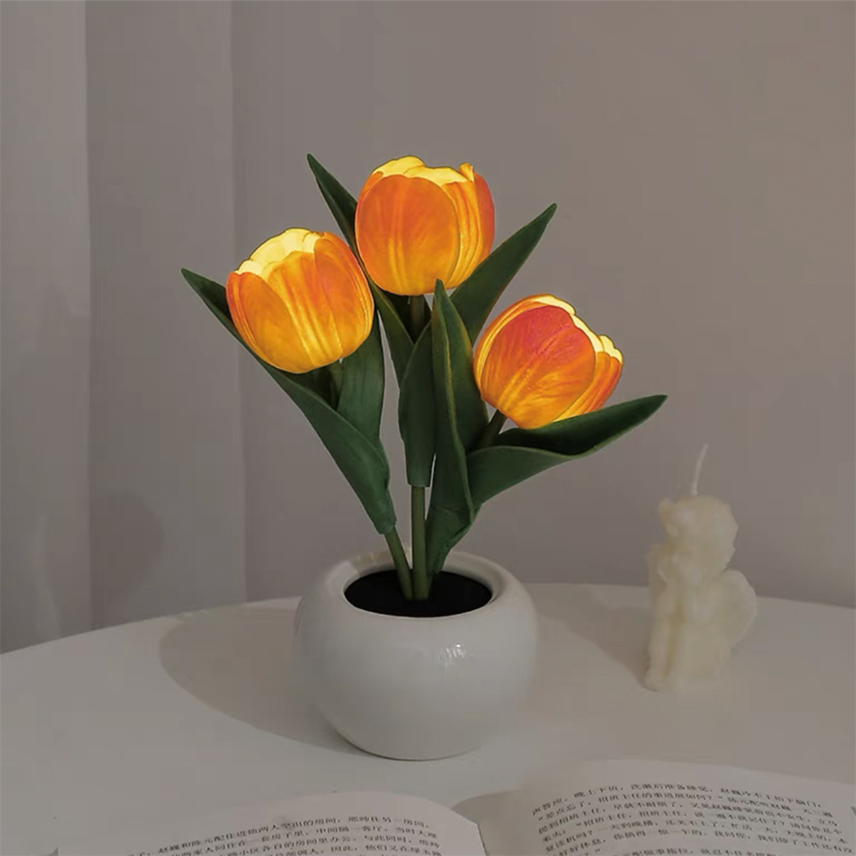Drei beleuchtete orangefarbene Tulpenblüten in einer weißen Vase auf einem weißen Tisch, mit Kerze, gedämpfte Umgebung.