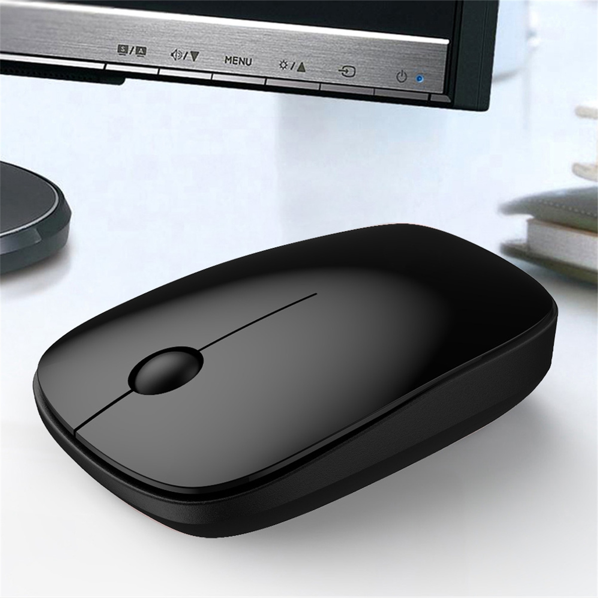Eine schwarze kabellose Maus auf einem weißen Schreibtisch. Ein Monitor ist im Hintergrund, unscharf.