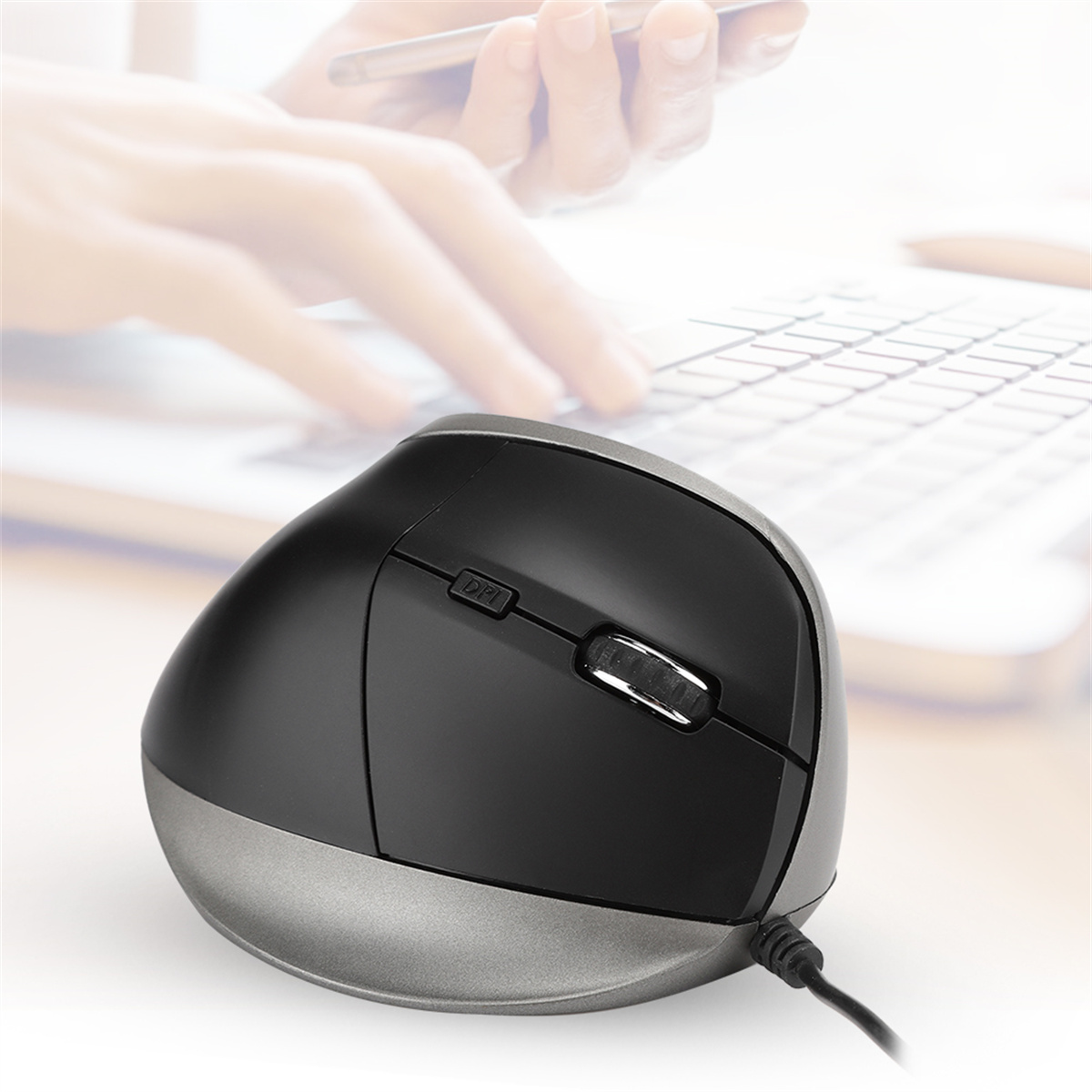 Schwarze und graue ergonomische Maus mit Kabel, in der Nähe einer Tastatur. Verschwommene Hände im Hintergrund.