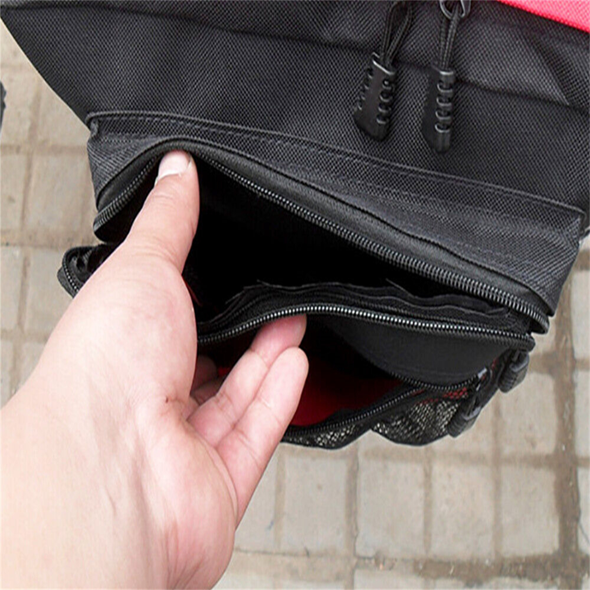 Hand öffnet eine schwarze Tasche mit mehreren Fächern und Reißverschlüssen, rotes Innenfutter sichtbar.
