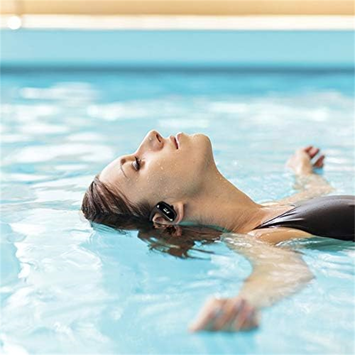 Frau im Wasser, mit Ohrhörern. Sie treibt, Augen geschlossen. Blauer Wasserhintergrund.
