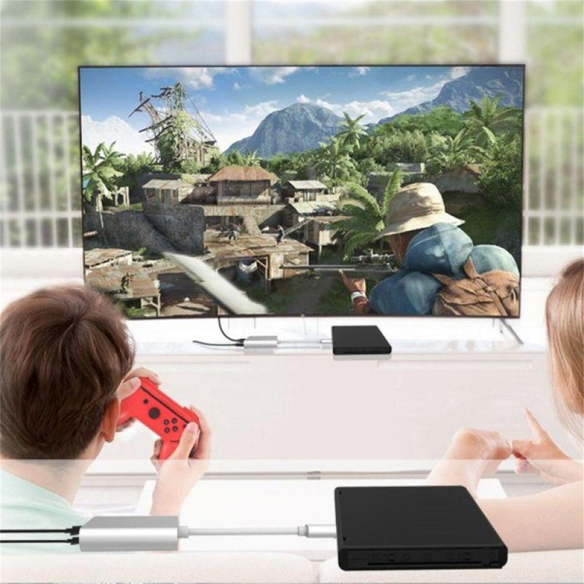 Zwei Personen spielen ein Spiel auf einem Fernseher. Einer hält einen roten Controller. Schwarze Boxen verbinden sich mit dem Fernseher.