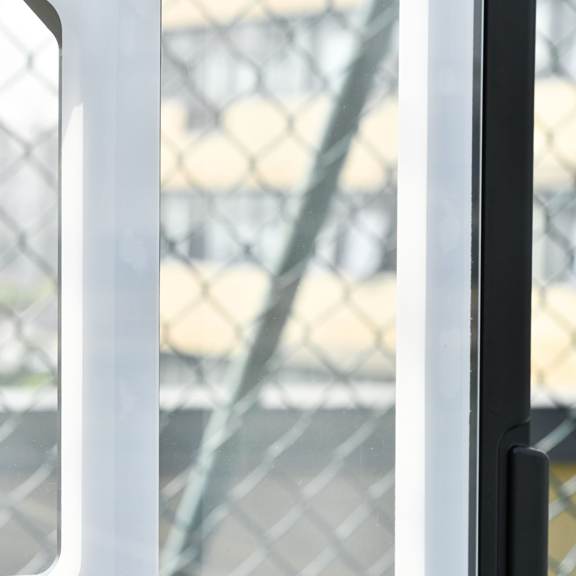 Ein Blick durch ein Fenster mit weißen Rahmen und einem schwarzen Griff. Draußen ist ein Maschendrahtzaun zu sehen.