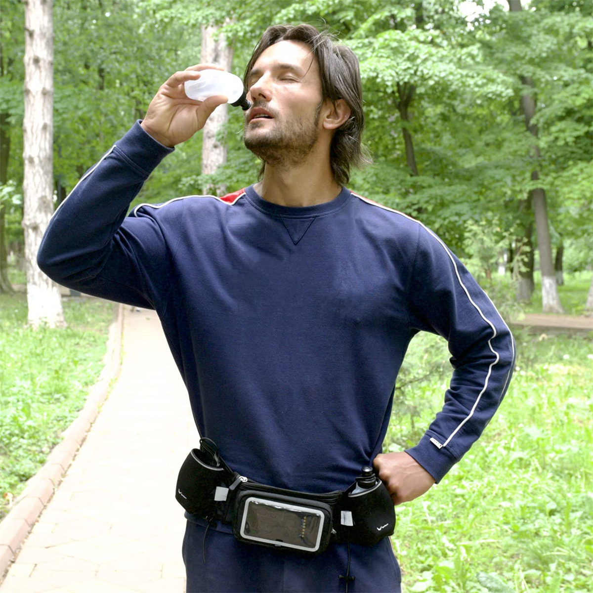 Ein Mann trinkt aus einer Wasserflasche, trägt ein blaues Hemd und eine Bauchtasche. Er ist in einem Park.