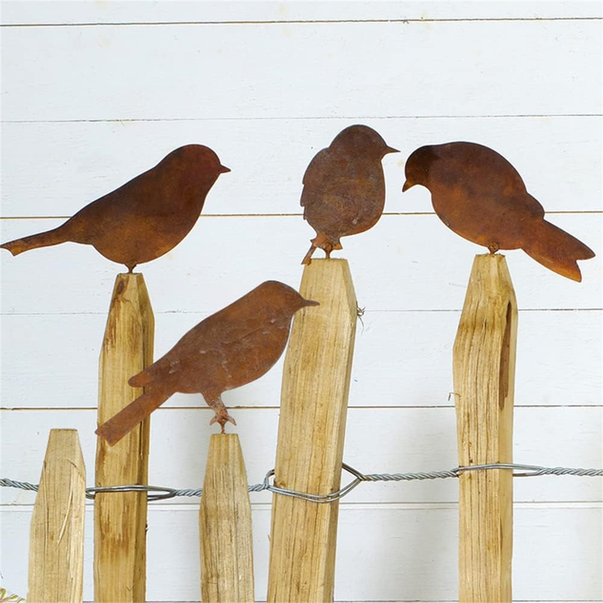 Vier rostige Metallvogel-Silhouetten auf Holzpfosten, vor weißem Holzgrund.