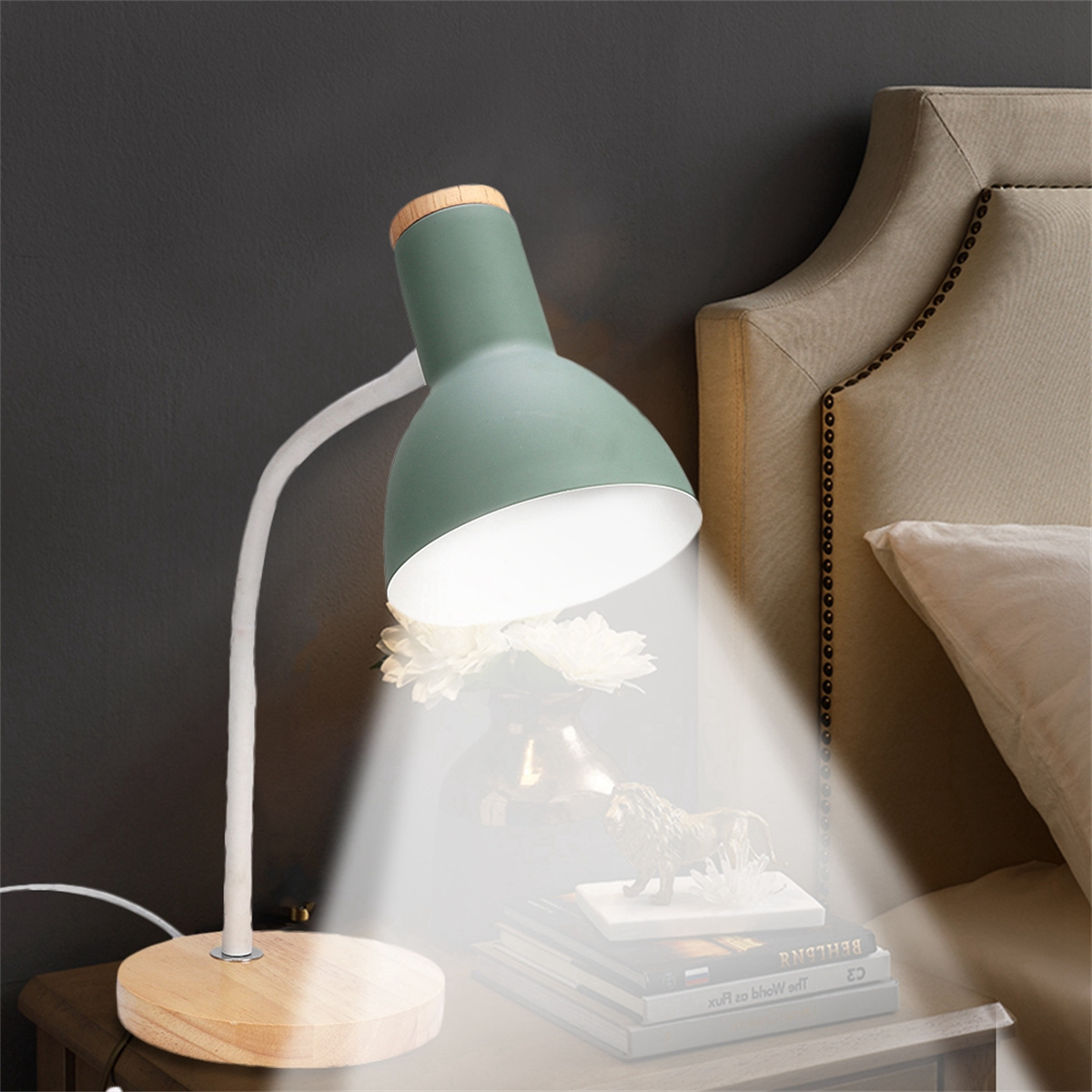 Eine grüne und hölzerne Schreibtischlampe beleuchtet Bücher und eine Figur auf einem Holztisch. Ein Bett im Hintergrund.