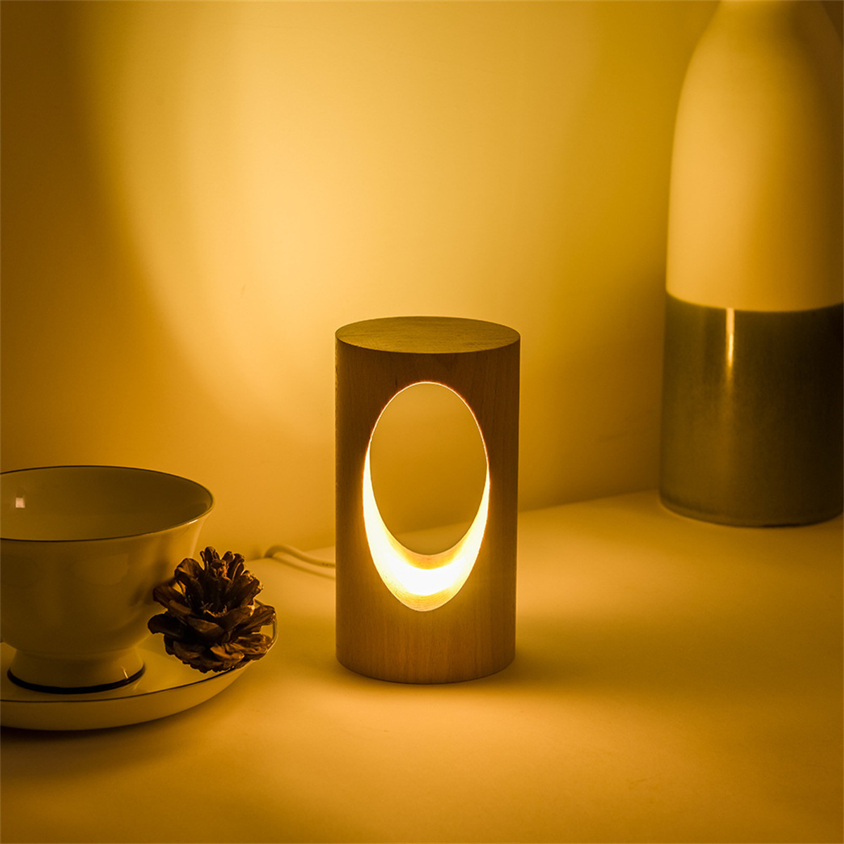 Holzzylinderlampe mit ovalem Ausschnitt, die warmes Licht aussendet. Eine Tasse und ein Tannenzapfen im Hintergrund.