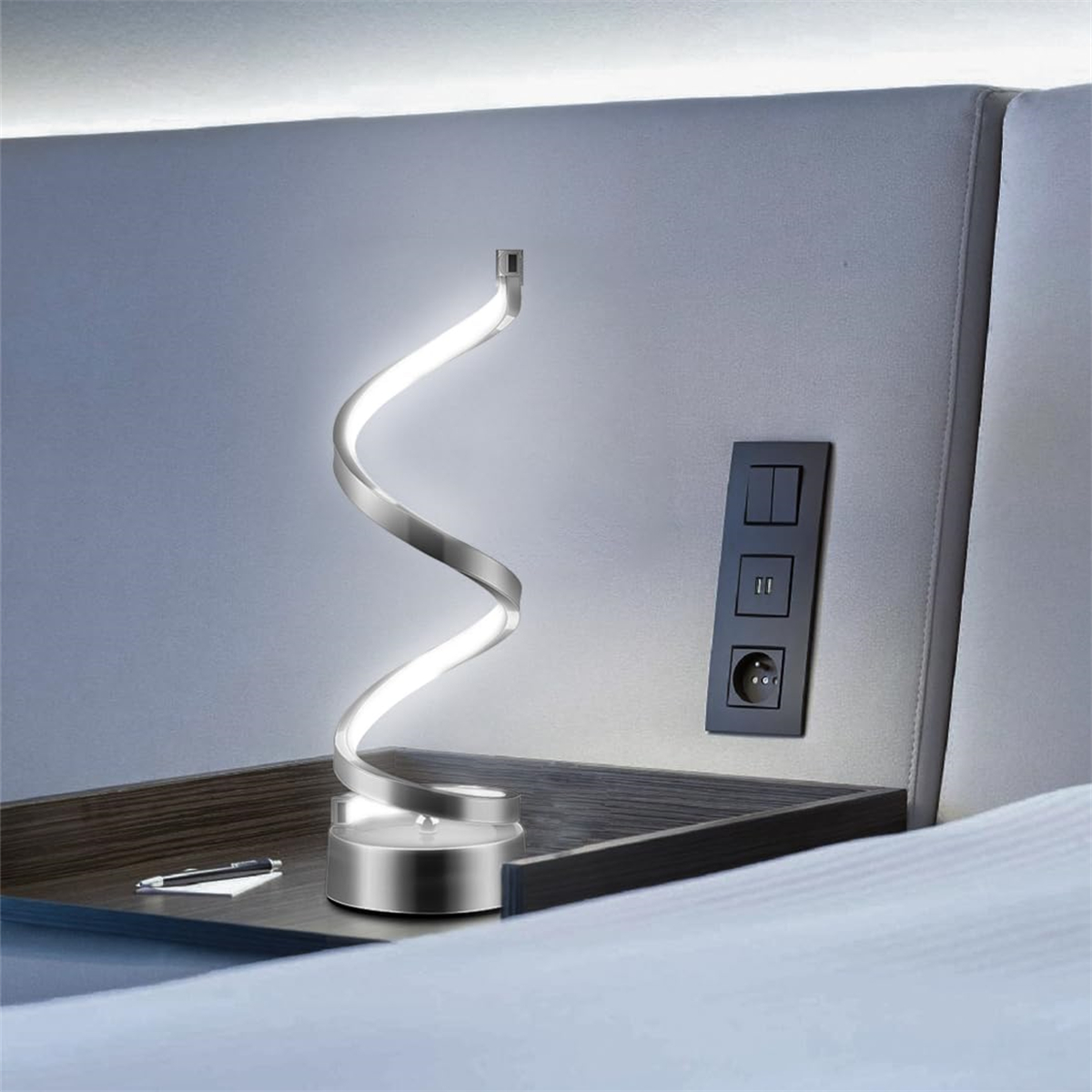 Silberne Lampe mit Spiraldesign auf einem Holztisch neben einer Steckdose.