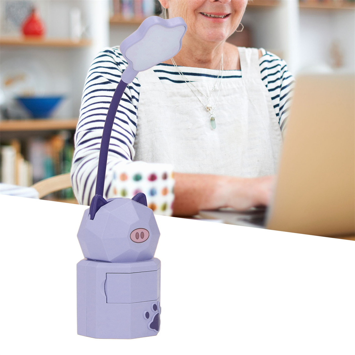 Eine lila, katzenförmige Lampe auf einem Schreibtisch, im Hintergrund eine lächelnde Frau am Laptop.