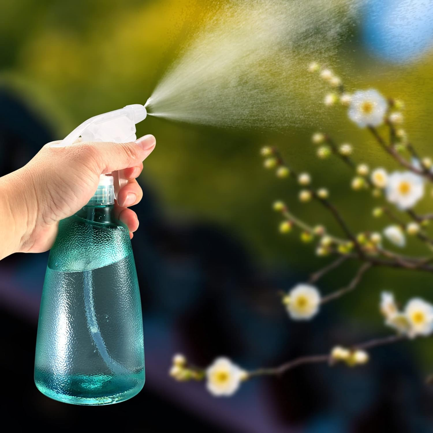 Eine Hand sprüht Wasser aus einer grünen Flasche. Weiße Blumen sind im unscharfen Hintergrund.
