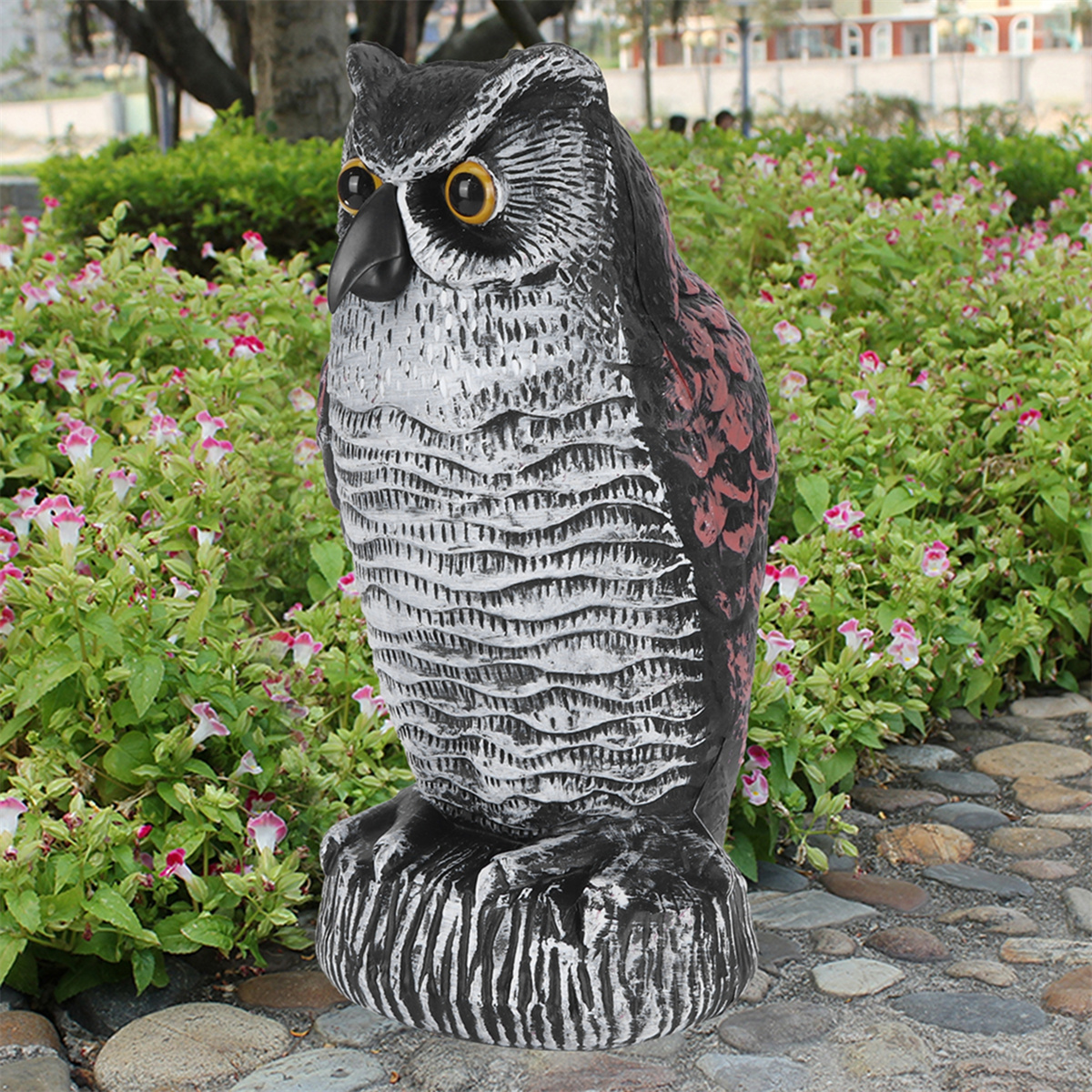 Eine Eulenstatue steht in einem Garten mit rosa Blumen. Sie ist schwarz, weiß und braun.