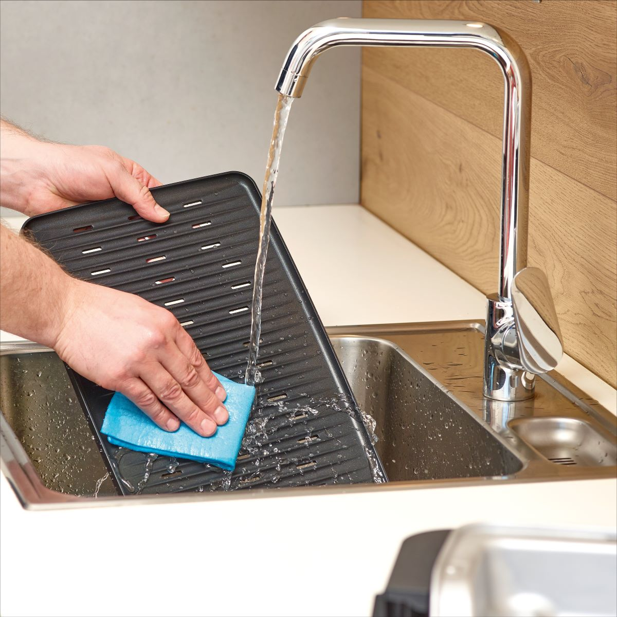 A person washing a black grill pan under a kitchen faucet with a blue sponge.