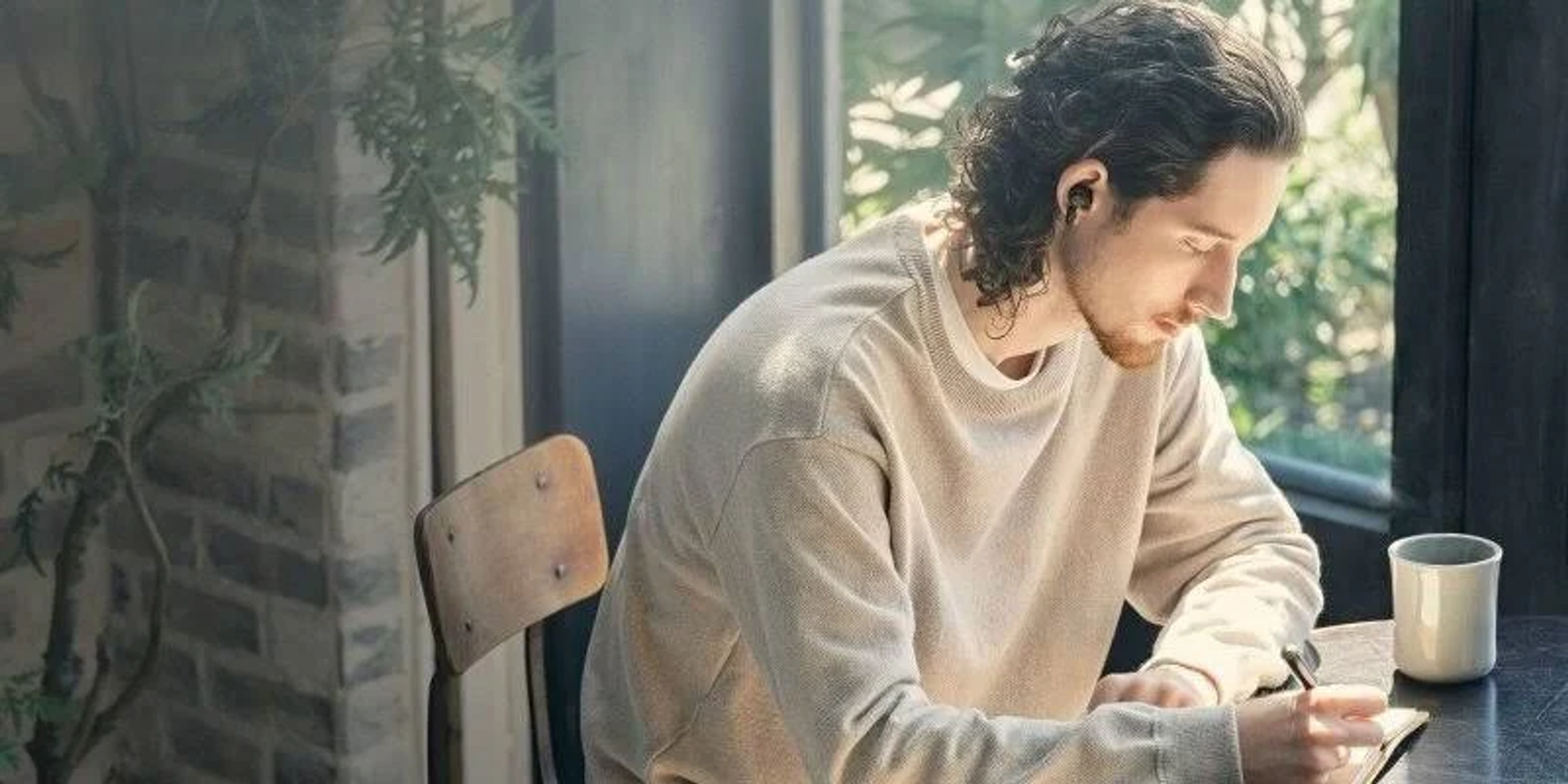 Mężczyzna w beżowym swetrze pisze przy stole, używając słuchawek.