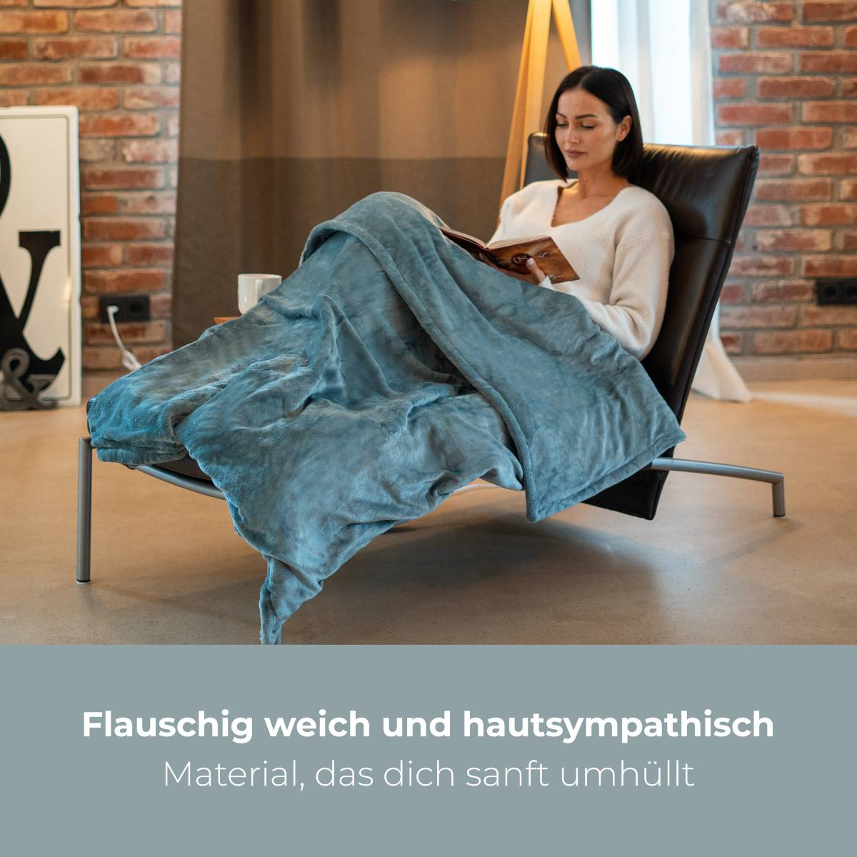 Kobieta siedzi na krześle czytając książkę, z niebieskim kocem. Tłem jest ceglana ściana i lampa.