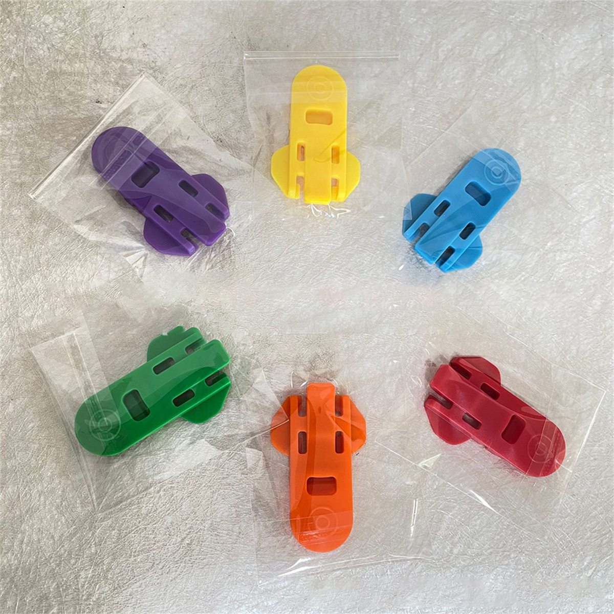 Sechs bunte Plastikclips in transparenten Beuteln. Jeder hat eine einzigartige Form und Farbe: lila, gelb, blau, grün, orange, rot.
