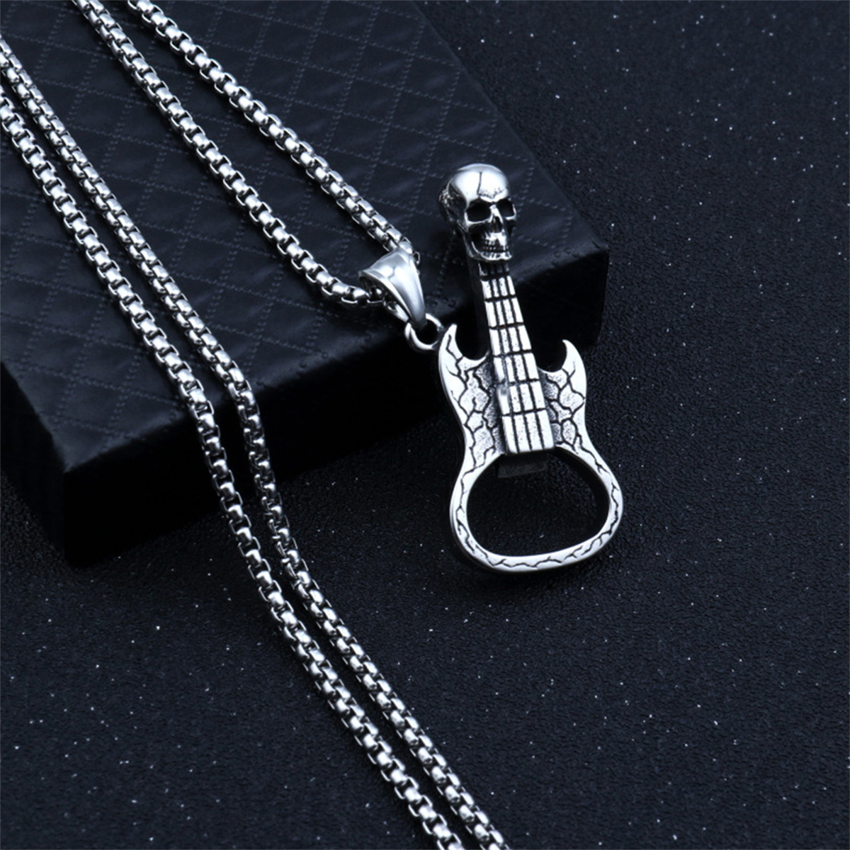 Eine silberne Gitarrenkette mit einem Totenkopfanhänger auf einer dunklen Schachtel. Die Kette liegt auf schwarzem Untergrund.