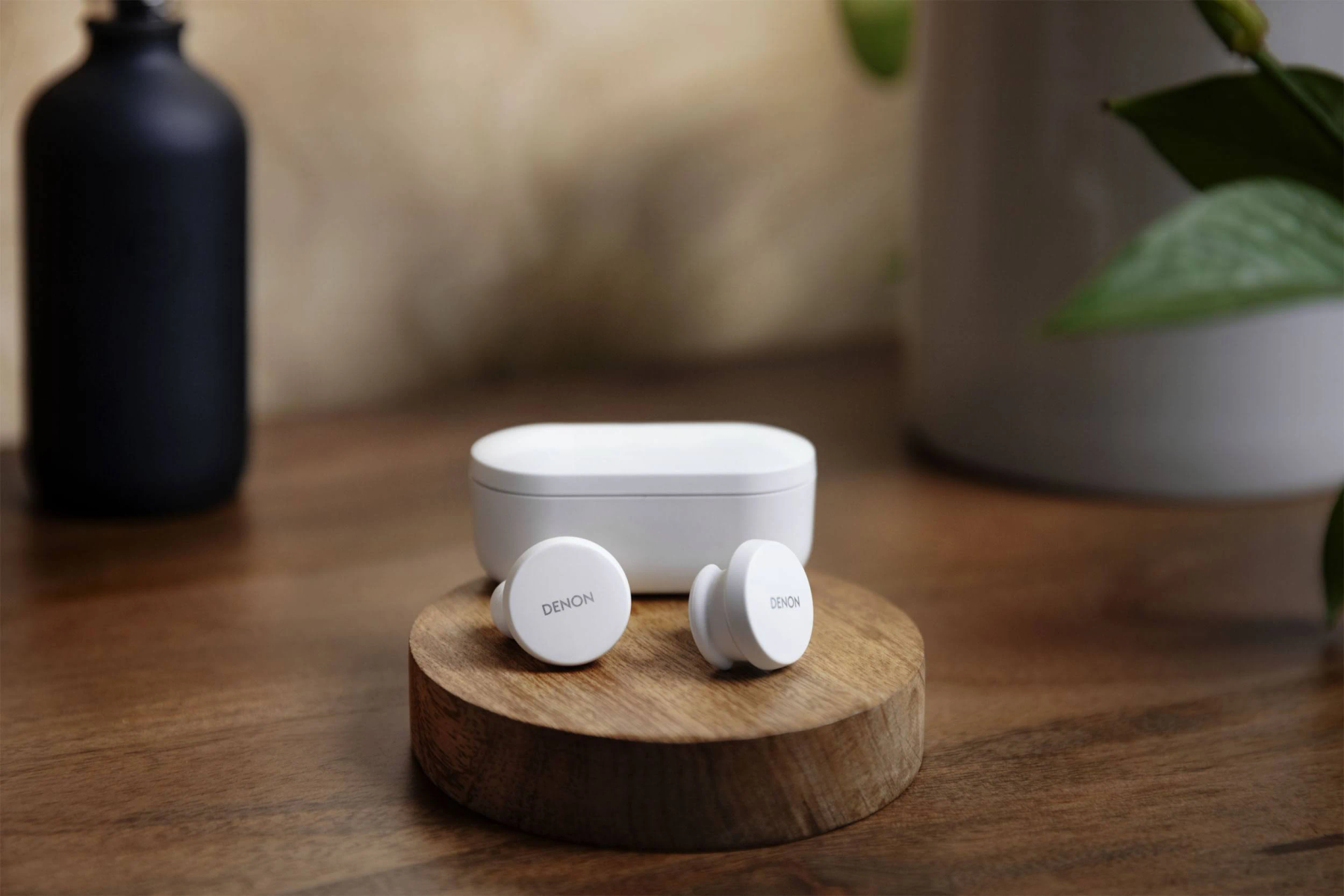 Auricolari bianchi e custodia di ricarica su un supporto in legno. Pianta e vaso nero sullo sfondo.