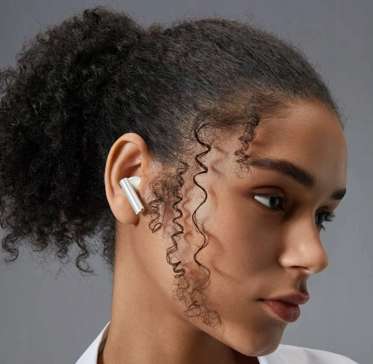 Donna con auricolare bianco nell'orecchio, capelli ricci, guarda a destra, sfondo grigio.
