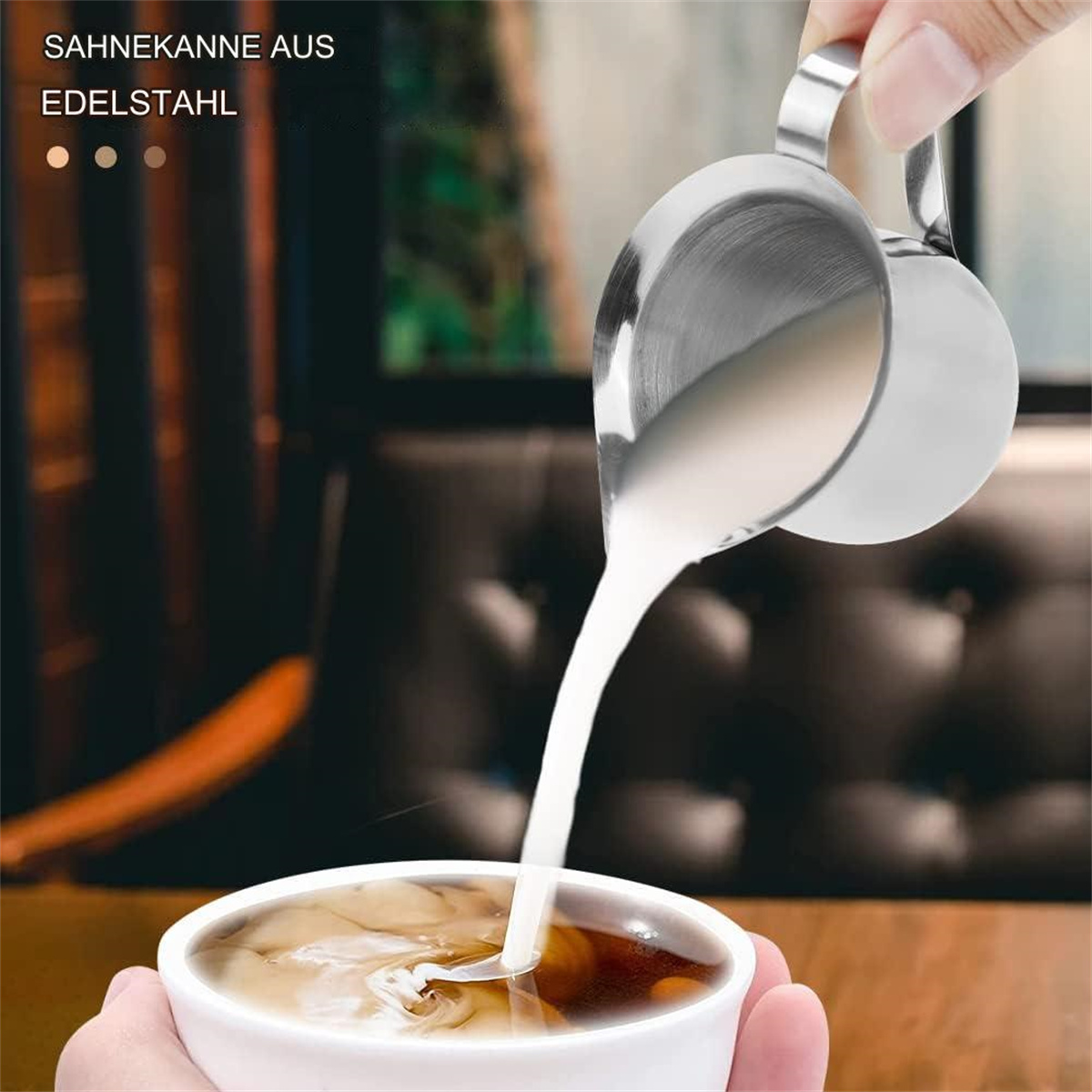 Silberne Milchkanne gießt Milch in eine Tasse Kaffee. Text: 'SAHNEKANNE AUS EDELSTAHL'.