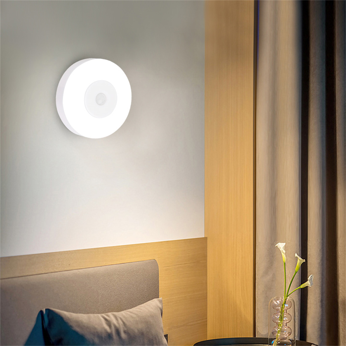 Eine weiße, kreisförmige Wandlampe mit Sensor, an einer grauen Wand neben einem Vorhang.