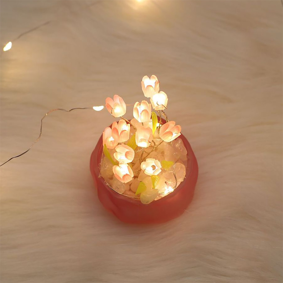 Kleiner roter Topf mit leuchtenden Blumenlichtern, auf weißer, flauschiger Oberfläche.