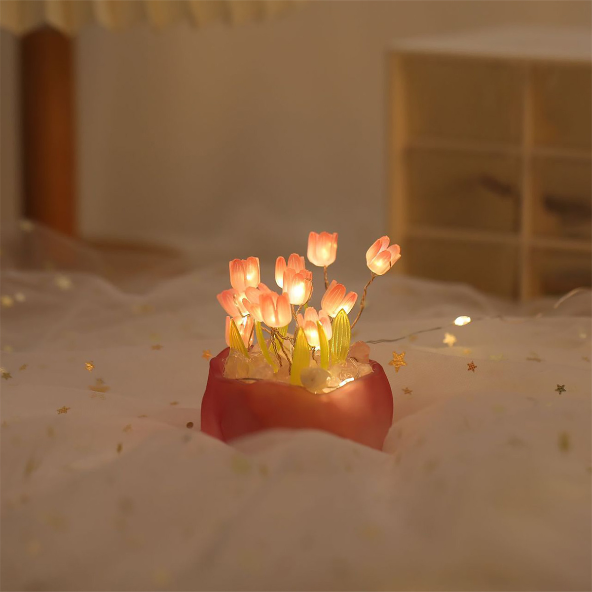 Kleine künstliche Blumenanordnung mit leuchtenden Lichtern, in einem roten Topf, auf einem weißen Tuch.
