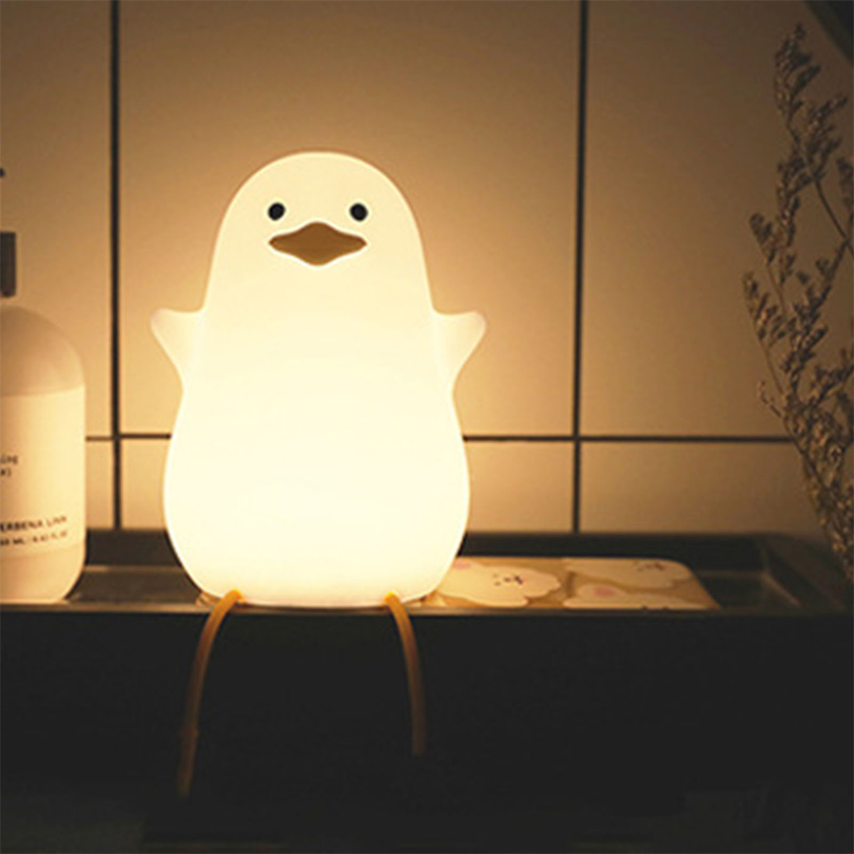Ein weißes pinguinförmiges Nachtlicht. Es ist beleuchtet, mit gelben Beinen, auf einem Regal sitzend.