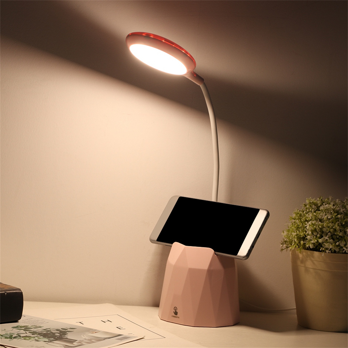 Eine Schreibtischlampe mit Telefonhalter und einer kleinen Pflanze auf einem Schreibtisch.